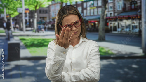 Billede på lærred Woman in white shirt and red glasses points finger forward with crossed arms on city street with buildings; accusation doubt confrontation