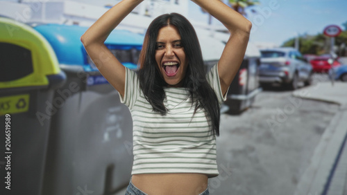 Fotografie Woman with arms raised above head beside large recycling bin, mouth open and tongue out in a spirited pose on a crowded urban street; playful joy