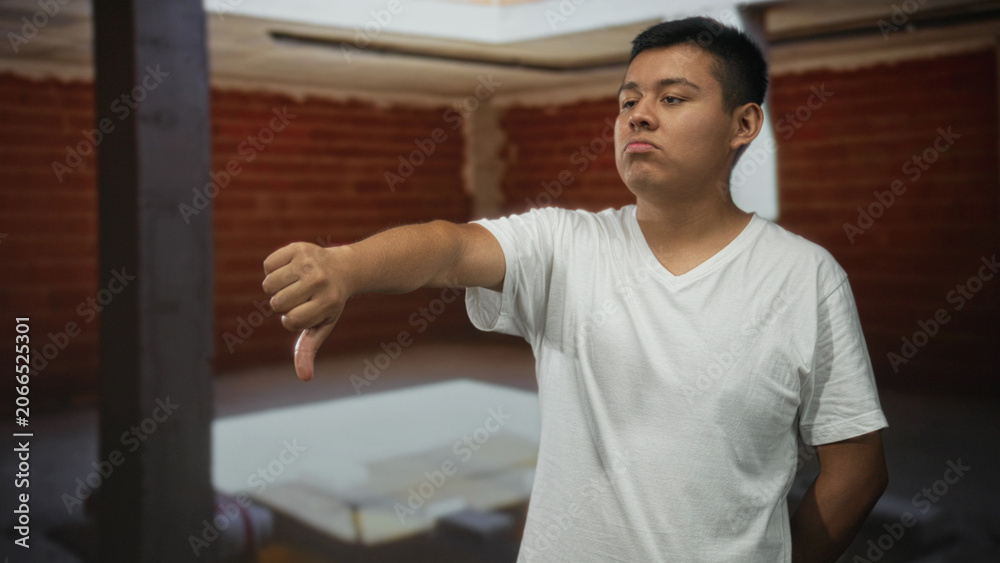 Fototapeta premium Man showing thumbs down with outstretched arm and visible thumb inside building interior with brick wall; disapproval critique.