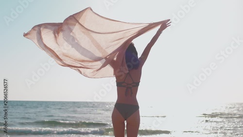 A woman in a bikini on a brightly lit beach, her arms raised and her scarf fluttering, creating a warm, sunlit silhouette. A sense of freedom and tranquility. 