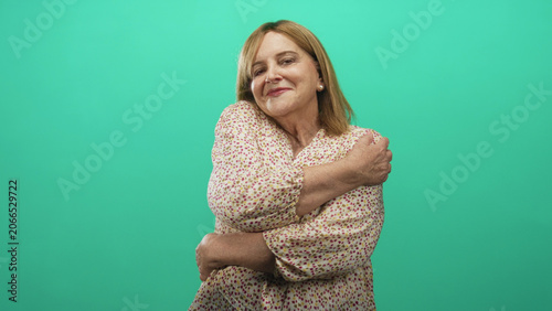 Obraz na płótnie Woman hugging her shoulders with bare hands and smiling in a studio against a teal backdrop, arms crossed in self embrace; contentment