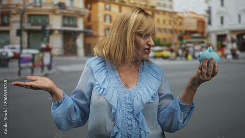 Photography Middle age woman holding blue piggy bank in right hand and gesturing palm up with left hand on a street near buildings, speaking to camera; saving concerns uncertainty