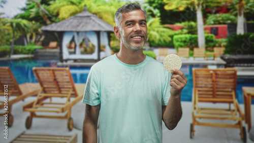 Fotografija Man holds ricecake in bare hand by resort building poolside cabana, smiling at camera; relaxation healthy snack
