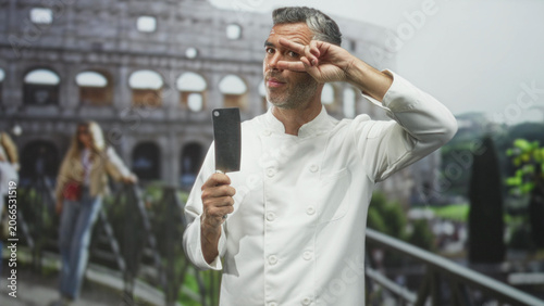 Obraz na plátně Grey haired man chef in white jacket holds cleaver in right hand and flashes v sign at eye in front of building colosseum; playful mischief