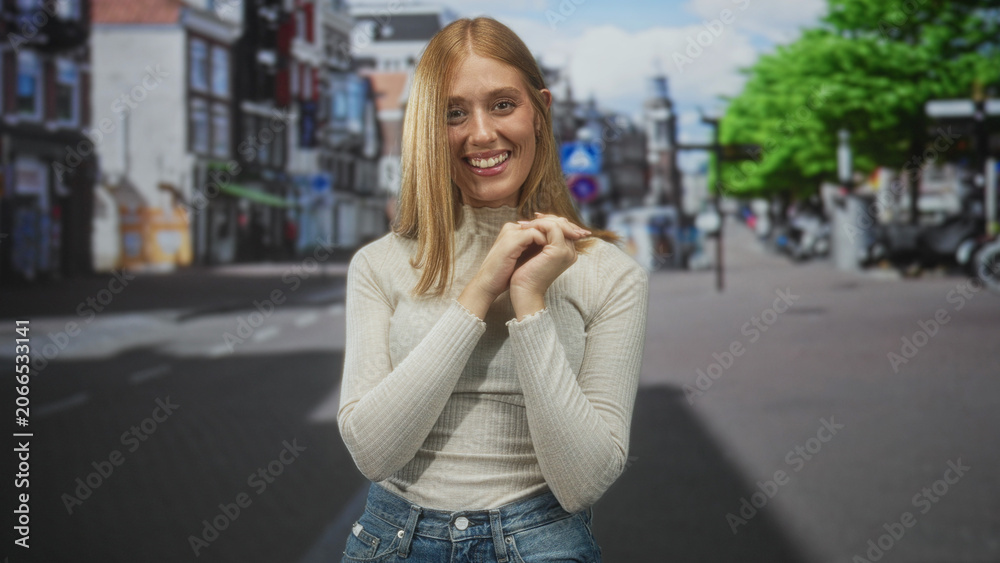 Fototapeta premium Redhead woman smiling and clasping hands near her face, wearing a turtleneck sweater and jeans while standing on a busy city street with storefronts; joy warmth.