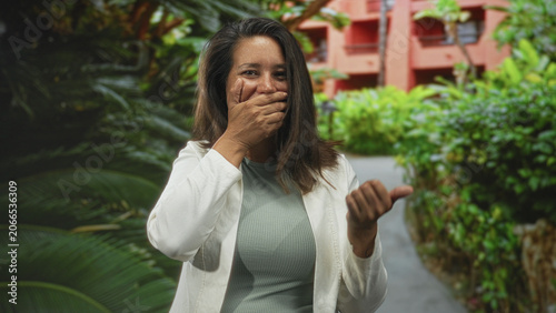 Valokuva Woman covers mouth with hand and points with other hand in building courtyard wearing white blazer and green shirt; surprise moment