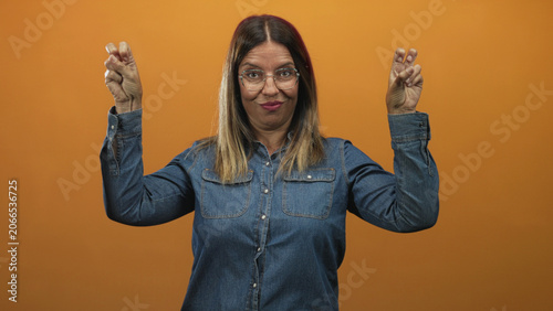 Fotografi Woman making air quotes with hands and smirking against an orange studio backdrop, wearing denim shirt and glasses; skepticism