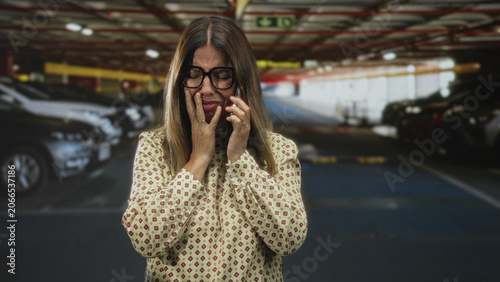 Cuadro en lienzo Woman wearing glasses holding phone, hand to mouth crying with fingers on cheek in parking building near cars and concrete pillars; anxiety urgency