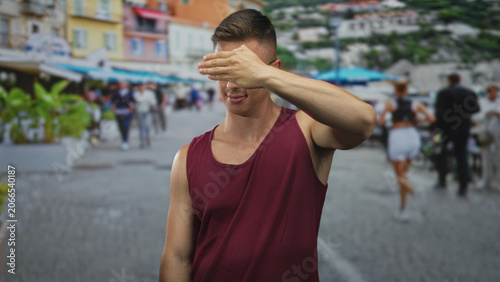 Canvastavla Man shielding eyes with hand while squinting, showing bare upper arm in a maroon tank top on a crowded cobblestone street with outdoor cafes; annoyance avoidance