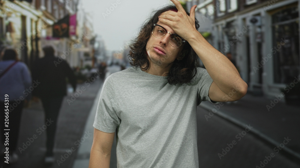Fototapeta premium Man, young with long hair and glasses, hand on forehead in a busy city street wearing gray t shirt; fatigue reflection.
