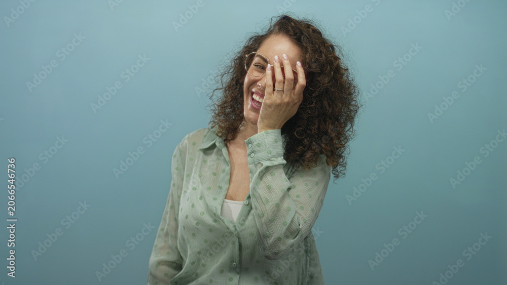 Fototapeta premium Woman laughing with hand covering face, wearing glasses and mint green polka dot shirt and curly hair in blue studio; joy authenticity warmth.