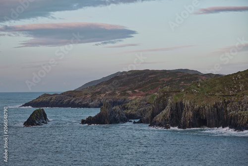 The ocean meets a rocky shoreline and sea stacks, with rolling hills covered in sparse vegetation forming the background.