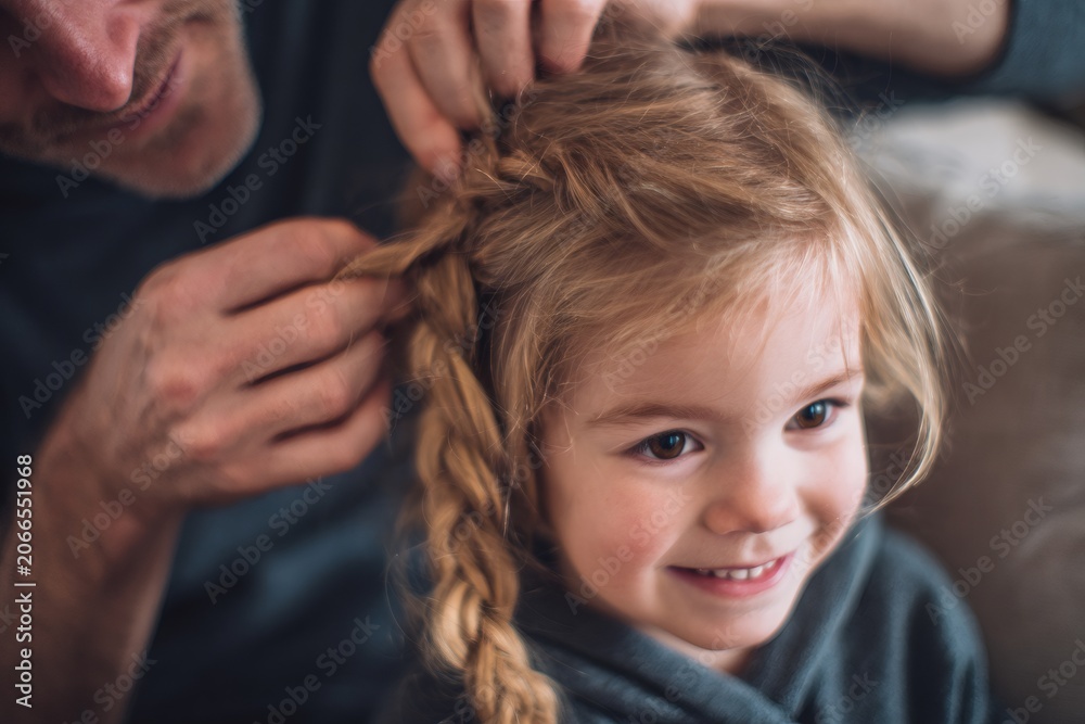 Naklejka premium Close-up of a father's hands braiding his little daughter's hair. Tender family moment at home. Paternal care and fatherhood concept