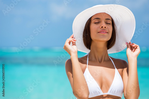 A woman stands on a sunlit beach, wearing a white bikini and large sun hat, basking in the warm sun with eyes closed, surrounded by blue ocean. Summer holidays concept. Generative AI.