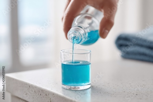 Person pouring blue mouthwash into a glass in the bathroom. Daily oral hygiene and dental care routine. Close up of fresh antiseptic liquid