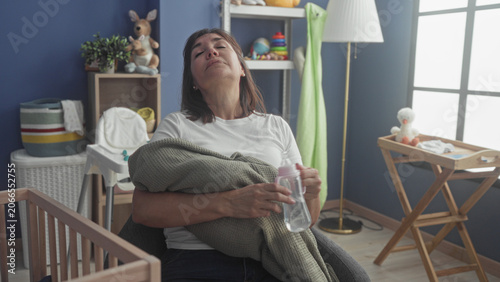 Bild auf Leinwand Woman cradles swaddled infant in green blanket, holds pink baby bottle and rubs forehead in nursery building; caregiver fatigue