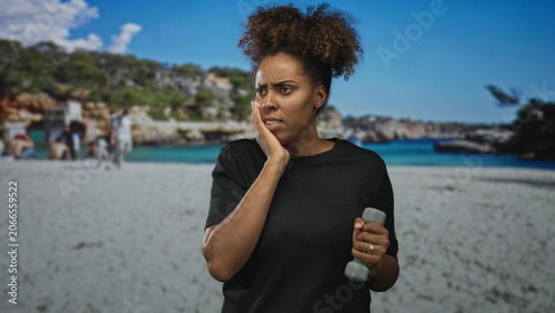 Fotografía Young african american woman grimacing with hand on cheek while holding a small dumbbell on a crowded sandy beach by the sea; pain toothache