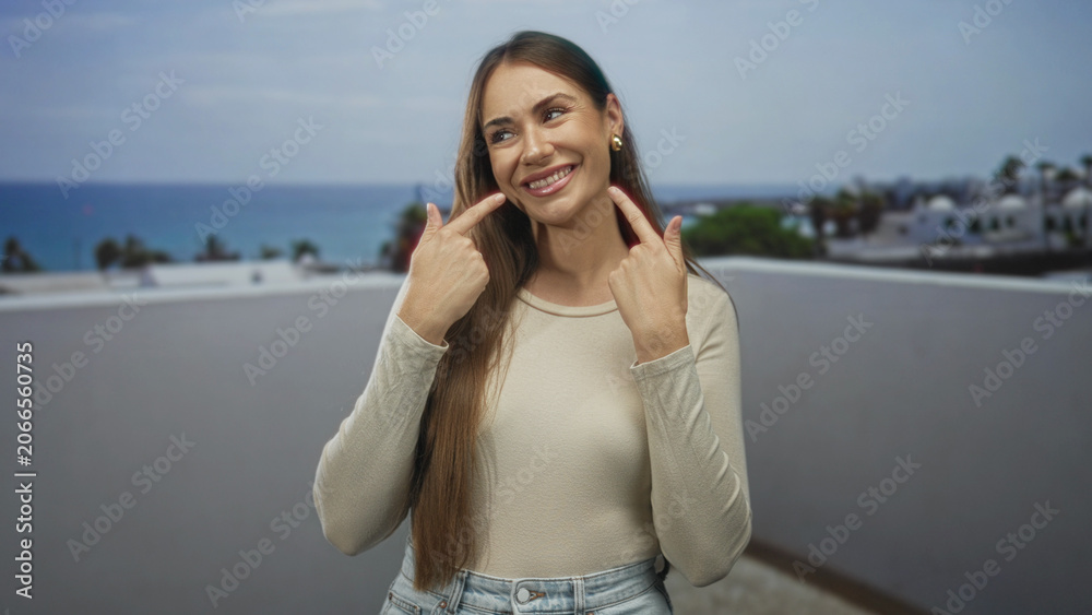 Fototapeta premium Young hispanic woman points index fingers to her smile showing teeth on a building terrace with ocean view; happiness confidence.