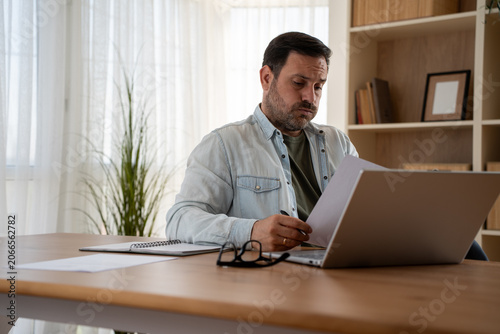 Worried man checking bills and financial papers while sitting at table with laptop. Stressed person planning home budget. Concept of personal finance review, expense control and household budgeting 