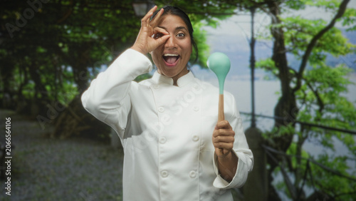 Foto Woman chef in white coat holding mint silicone ladle, smiling with open mouth and making ok gesture to eye at waterfront, trees and railing visible; playful creativity