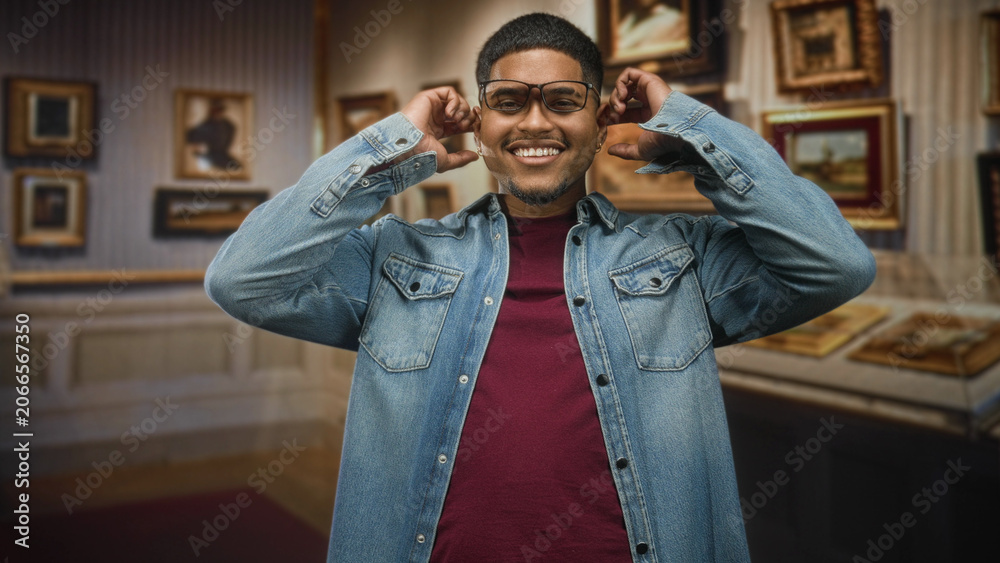 Fototapeta premium Hispanic man adjusts glasses and smiles while touching ears amid framed paintings in an art gallery building; joy wonder.