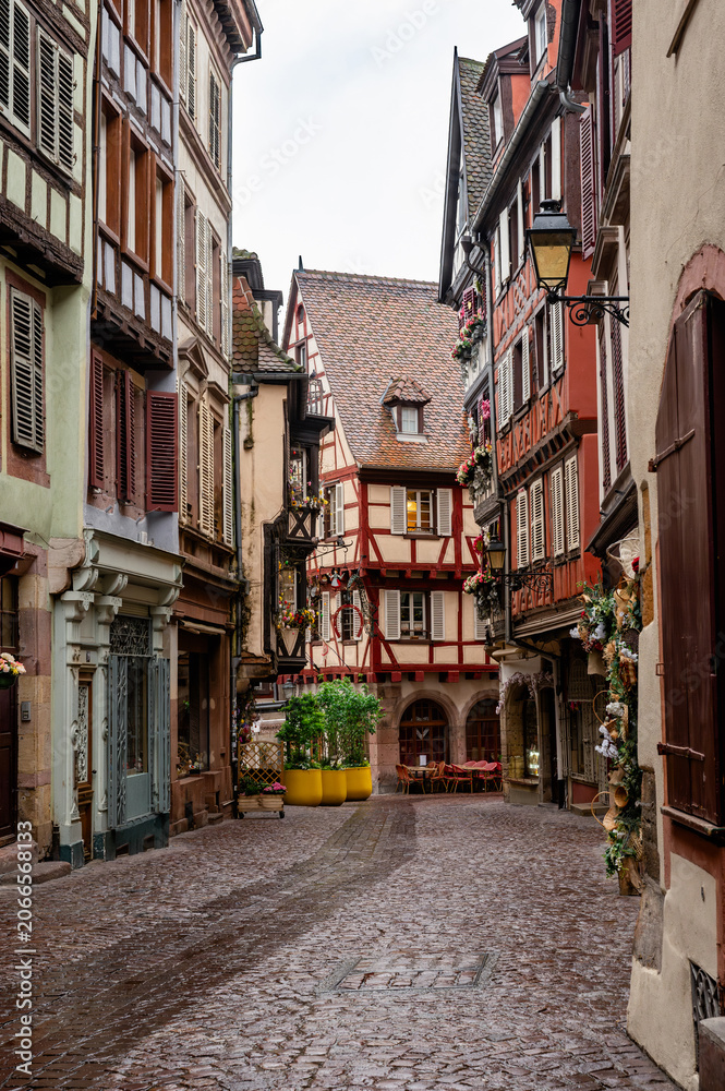 Fototapeta premium Intimate shuttered alley framed by red timbered facades, glossy cobbles, small planters and warm shopfronts, quiet rainy day mood ideal
