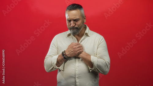 Fotografie Man clutching chest with closed eyes, middle aged hispanic with grey beard and bracelets, wearing light shirt in red studio; remorse reflection