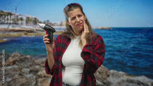 Woman holding cordless screwdriver, hand on cheek, red plaid shirt on rocky seaside shore with visible waves and rocks; toothache concern.