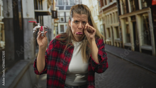 Canvas Print Woman holding metal faucet, rubbing eye and pouting on a cobbled street; plumbing problem frustration