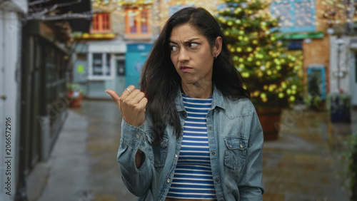 Fotografía Woman pointing thumb over shoulder on a wet street with storefronts and decorative lights; frustration
