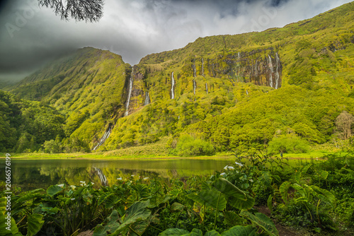 Fotografie Impresionante vista panorámica de cascadas cayendo por un acantilado verde, reflejadas en un lago rodeado de densa vegetación, Flores, Azores