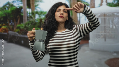 Tableau sur toile Woman points finger while holding a stovetop coffeepot and wearing a black and white striped shirt on a street with palm trees and a white gazebo; urban defiance