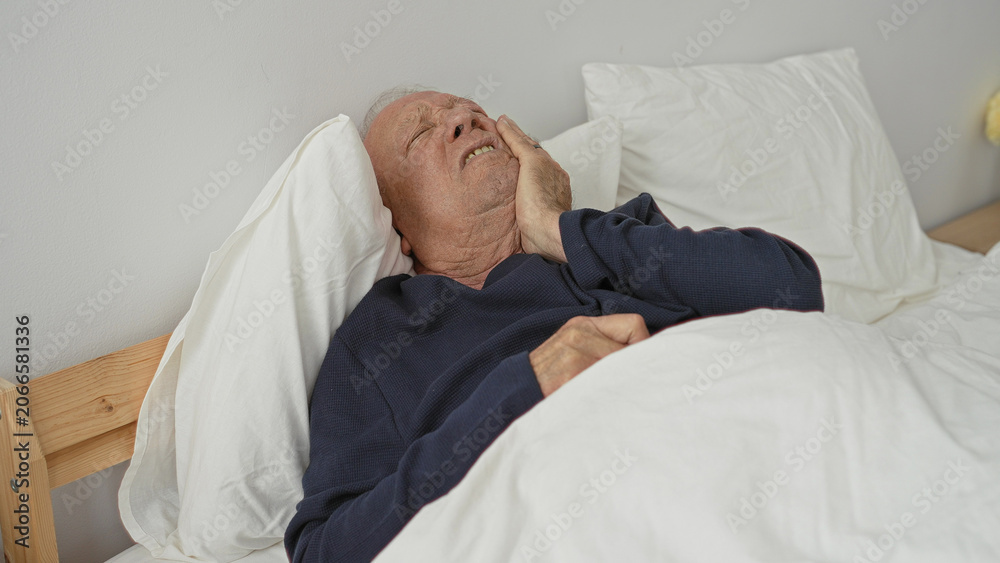 Fototapeta premium Grey haired elderly man lying in bed with hand on cheek clutching toothache, wearing navy top with pillows and duvet visible in a residential building; pain concern.