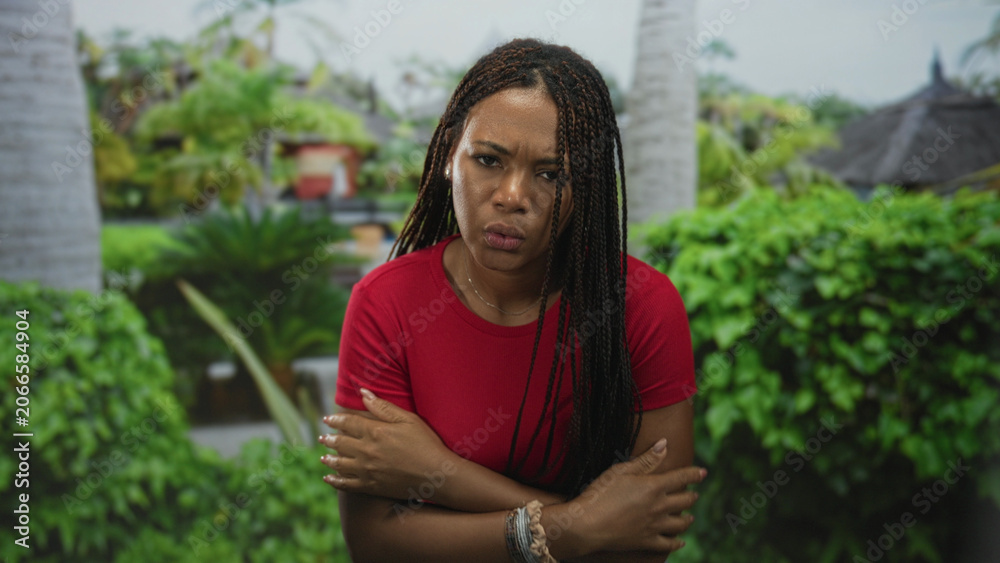 Fototapeta premium Woman with hands to mouth and arms crossed in park near palm tree, wearing red top and bracelets; discomfort cold.