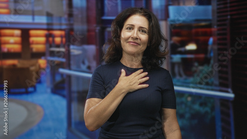 Photos Middle aged hispanic woman with hand on chest gesture in building, navy shirt, soft smile, looking at camera, relaxed posture; gratitude sincerity warmth