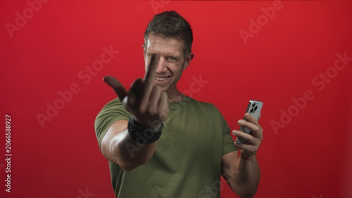 Obraz na plátně Man holding smartphone and extending middle finger toward camera in a red studio with wrist bracelets; anger defiance