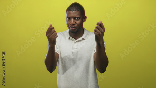 Photos Young black man with pinched fingers gesture and visible fingers aimed at camera in studio against yellow backdrop, wearing white polo; wry skepticism