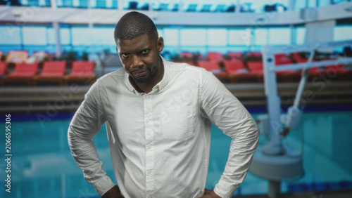 Photos Man in white shirt standing with hands on hips at an airport terminal beside rows of red seats and large windows, brow furrowed and head tilted; impatience doubt
