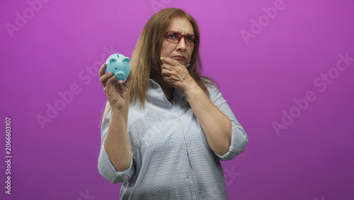Photography Senior hispanic woman holds blue piggy bank with hand on chin thinking in studio; financial worry savings