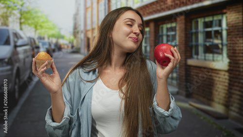 Fotografija Woman holding red apple and muffin in hands on street near parked cars and brick building, eyes closed deciding between snacks; choice contentment