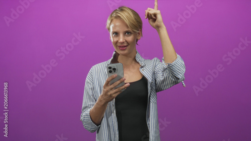 Canvastavla Blond woman holding smartphone, raising index finger and glancing aside, wearing striped shirt over black tank top in studio; confidence instruction