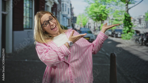 Obraz na płótnie Woman in pink striped shirt and glasses gestures with open palms toward street, showing smartwatch and bracelet; playful confidence