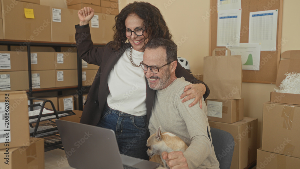 Fototapeta premium Man holding small dog and high fives woman at laptop amid parcel boxes in packing building; teamwork joy entrepreneurship.