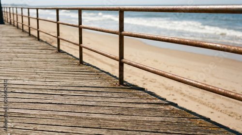 Old weathered wooden boardwalk with rusty metal railing extending towards the ocean. Calm waves breaking on the sunny sand