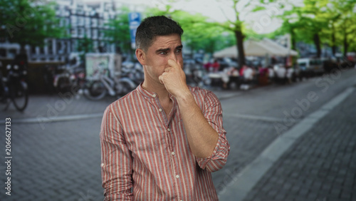 Photos Man pinching his nose on a busy city street beside parked bicycles and outdoor cafe seating, reacting to a foul smell; public disgust