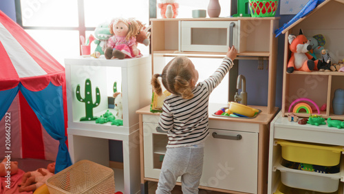 Foto Girl holds toy sandwich and reaches for a toy microwave while playing at a pretend kitchen in a building playroom; playful bonding