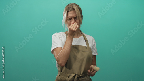 Photos Woman makeup artist young blonde wearing apron pinches nose while holding brush in studio with teal backdrop; disgust reaction