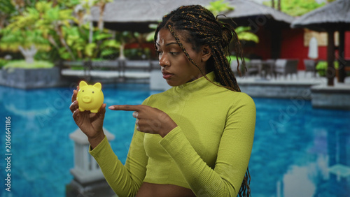 Konstfotografi Woman points finger at yellow piggybank by hotel pool in resort building, holding piggybank with bare hand; saving money thoughtful