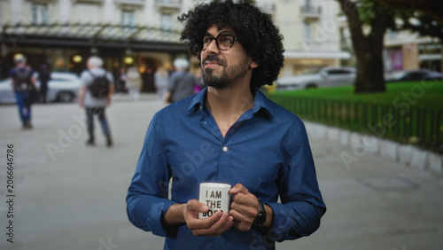Fotografía Man holding mug with both hands on street near park fence wearing blue shirt and round glasses; contentment quiet focus moment