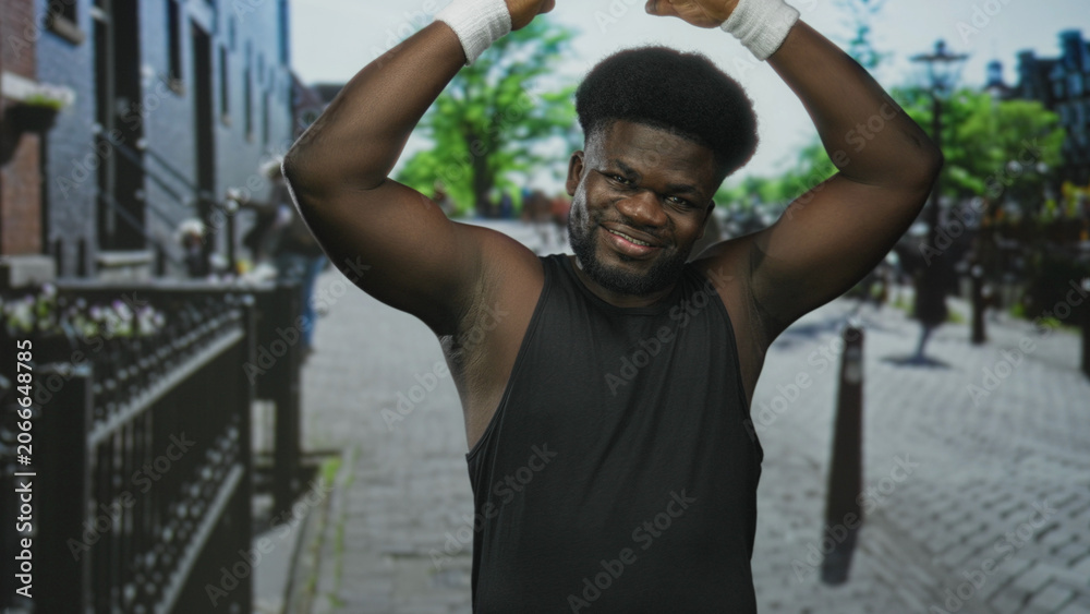 Fototapeta premium Young african american man raising bare arms overhead on city street in black tank top with white wristbands and smile; confidence fitness motion.